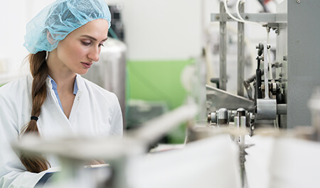 Machinery Industry - Happy female employee wearing protective headwear and white lab coat while working as a manufacturing engineer in a contemporary factory